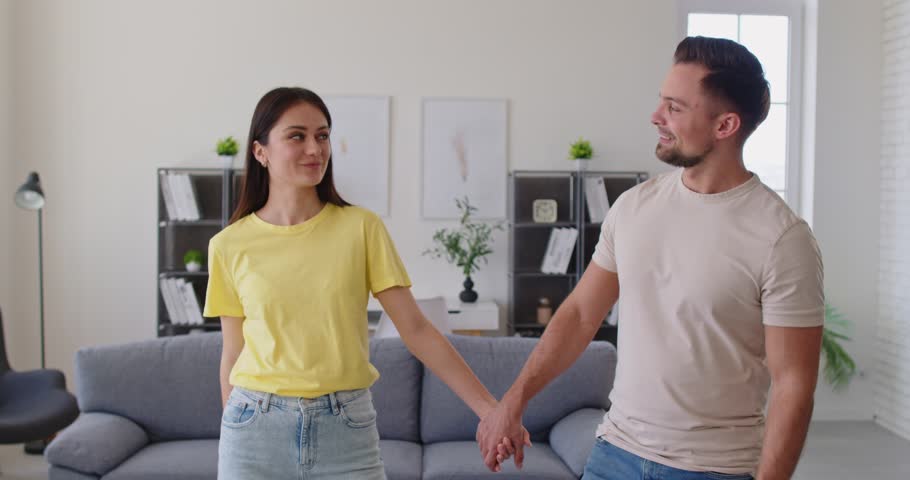 Young couple holding hands, standing in modern living room, looking out through windows, admiring view from their purchased or rented house. First homeownership, starting new chapter in life together
