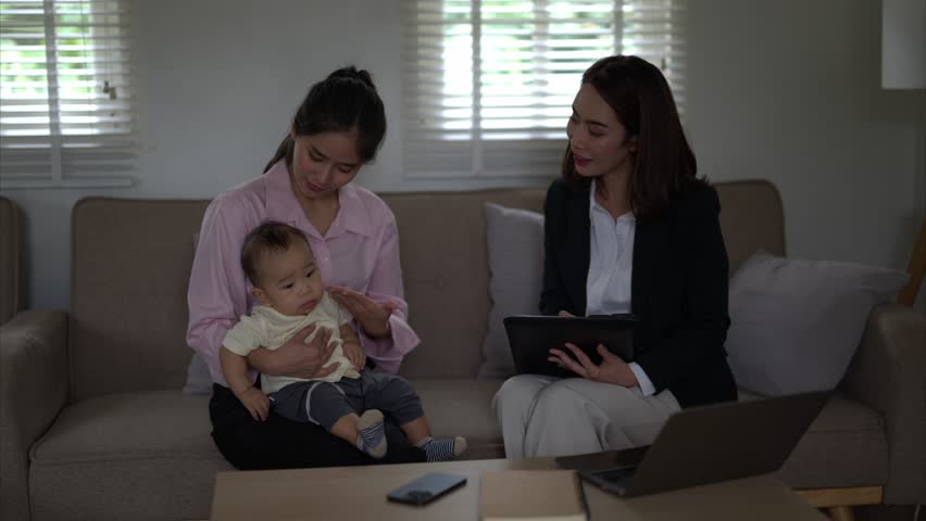 A woman is holding a baby on a couch while another woman sits behind her. The woman holding the baby is wearing a pink shirt