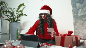 Young woman in Santa hat shopping online for Christmas gifts using tablet and credit card, sitting in cozy decorated room with lights and presents. - Powered by Shutterstock - Get 15% off with code: PIKWIZARD15