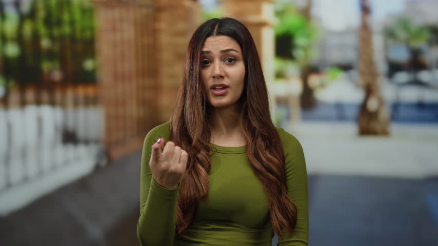 Woman gesturing no with finger in urban street setting, wearing green top on a sunny day, conveying refusal or caution outdoors, highlighting hispanic heritage and youthful appearance.