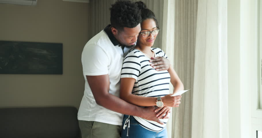 Couple noticing ultrasound printout at living room window embracing bump celebrating pregnancy. Romance, intimacy, family, maternity, joy, tenderness, connection