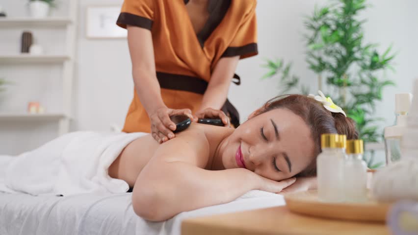 Beautiful relaxing woman getting back massage therapy with hot stone. Attractive young girl lying on massage bed, getting physiotherapy from masseuse for skin and body care in spa beauty clinic.
