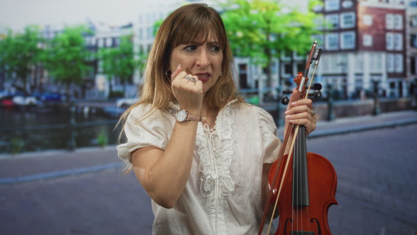 Young woman bites fingernail while holding violin and bow on busy city street by canal with brick buildings and trees in daylight; stage anxiety performance nerves.