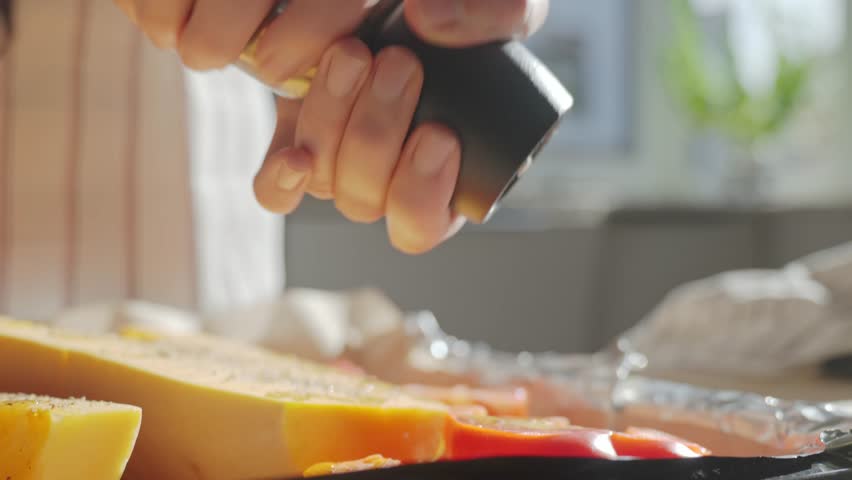 Person sprinkling butternut squash and vegetables at kitchen. Hands using pepper grinder to season fresh pumpkin and vegetables on baking tray. Concept of home cooking and healthy food preparation