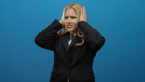 Woman in business suit covering ears against blue background, expressing stress and annoyance with closed eyes, emphasizing frustration through body language and facial expression. - Powered by Shutterstock - Get 15% off with code: PIKWIZARD15