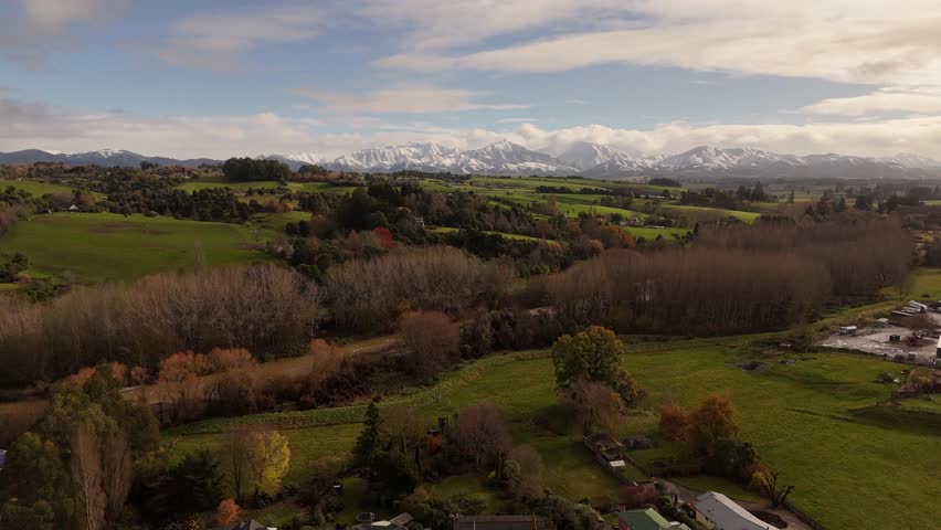 River surrounded by withered trees near green hills with snow-covered mountains during autumn, aerial shot
