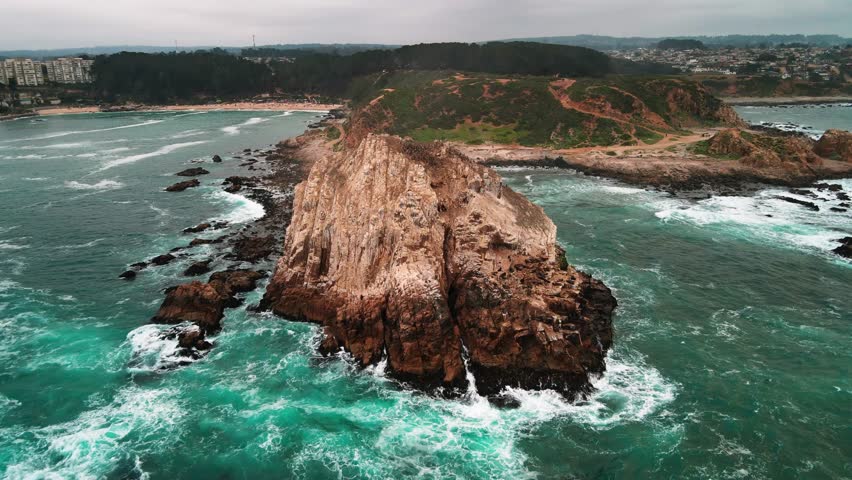 Drone aerial forward over a rocky coastal headland and bay in the afternoon, capturing rugged cliffs, surf and seabird rookeries in moody overcast light.