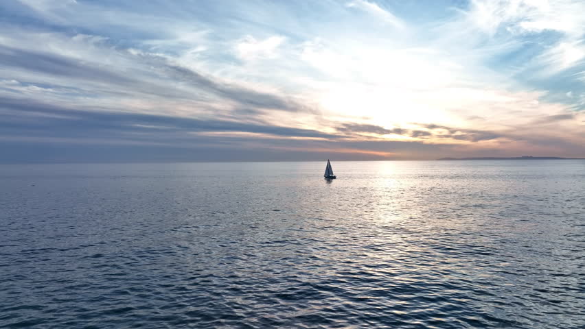 A sailboat moves across the sea. The boat sails on sea. Sailboat floats in open water. The sail catches the evening wind. The sailboat drifts past the glowing line.
