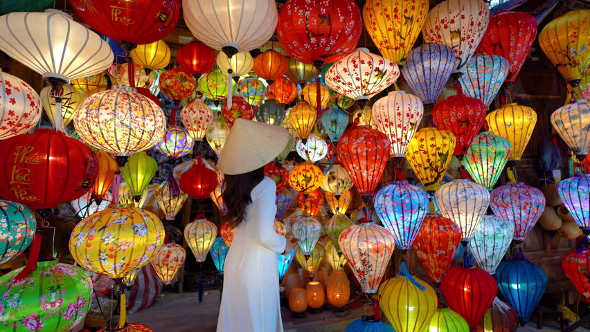 Beautiful Asian woman wearing traditional white ao dai dress with colorful paper lanterns in Hoi An, Vietnam.