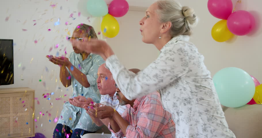 Diverse senior friends blowing confetti into air, celebrating at home and dancing under balloons. Celebration, friendship, joy, vitality, merriment, festive, lively