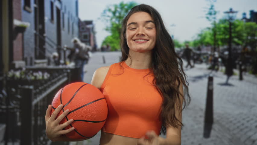 Young hispanic woman smiling and holding basketball and giving thumbs up on street; confidence encouragement.