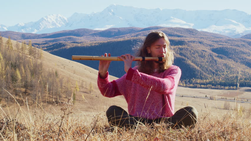 Serene musician with long hair sitting cross-legged in the dry grass, playing a traditional wooden flute against a breathtaking backdrop of majestic, snow-capped mountains on a sunny day