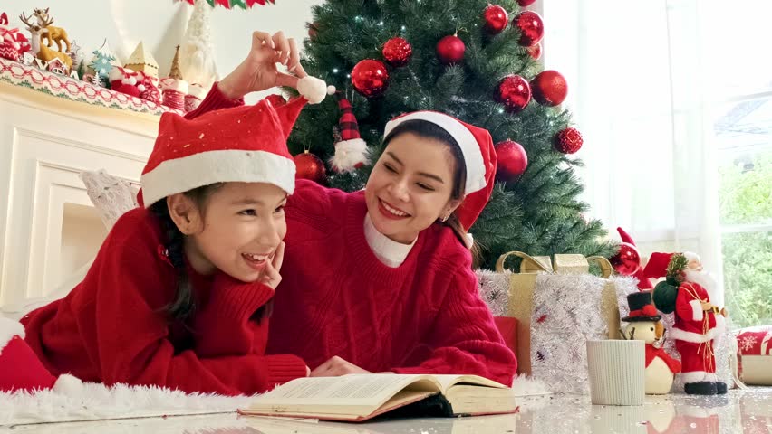 Asian mother and daughter child in Santa hats laugh and read on the floor beside a decorated Christmas tree, surrounded by gifts and cozy holiday decor in a warm living room. Real time 4k video