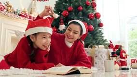 Asian mother and daughter child in Santa hats laugh and read on the floor beside a decorated Christmas tree, surrounded by gifts and cozy holiday decor in a warm living room. Real time 4k video - Powered by Shutterstock - Get 15% off with code: PIKWIZARD15