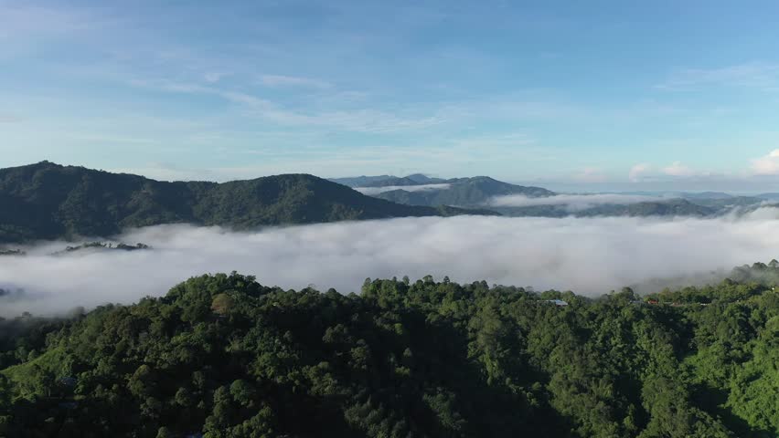 Aerial view of morning mist covering lush green mountains at Tamparuli, Sabah, Malaysia. Stunning tropical highland landscape with fog rolling over rainforest hills under clear blue sky.