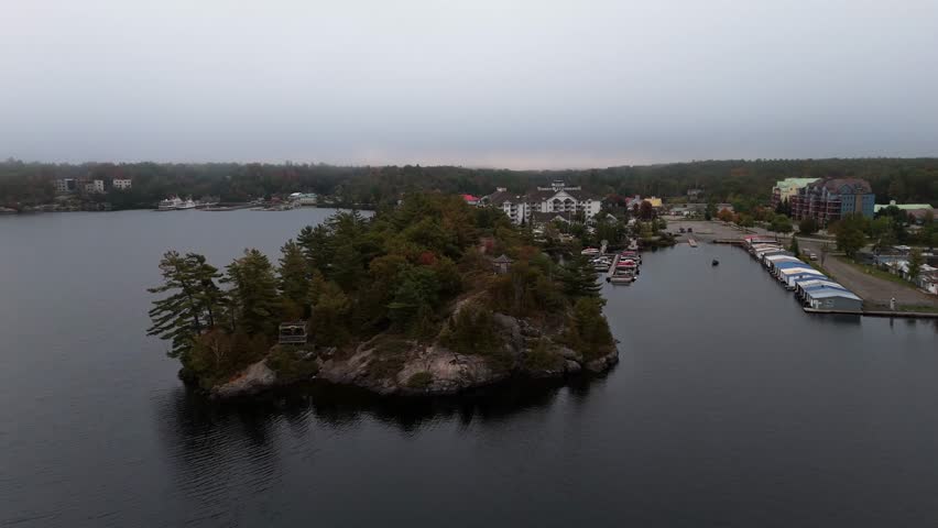 An aerial view of a quaint harbor and colorful boathouses along the wooded shoreline of Muskoka Wharf on Lake Muskoka with foggy background in Canada