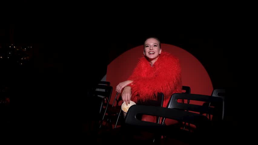 Portrait of a beautiful young woman in a red fur coat against a red background, posing for a cinema-style photo. The woman smiles beautifully, sitting on chairs in the light of a projector during a