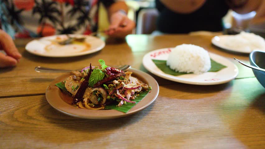 Person enjoys nam tok moo spicy pork salad with rice at wooden table, natural daylight.