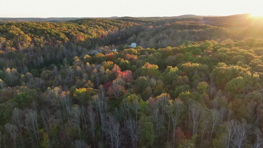 Aerial wide shot of river between colored trees in fall season. Sunset behind mountains in idyllic woodland of America. Peaceful scene.