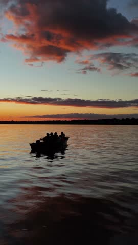 A vertical drone footage of a silhouetted boat with people cruising along the Amazon River at sunset, with dramatic clouds reflecting on the water.