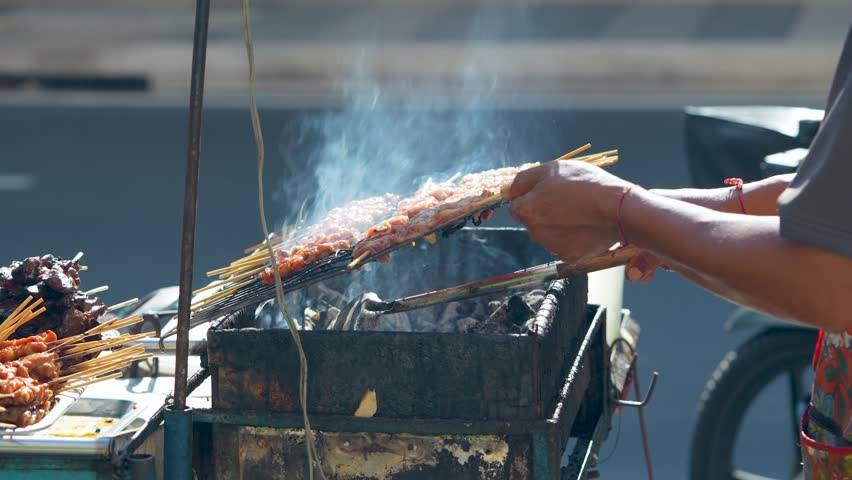 Vendor grilling chicken on sticks on homemade grill for customers outdoors at street food market in south Asia. Person cooking and selling unhealthy snacks, junk food at local bazaar in urban city.