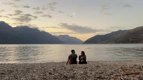 A couple sits together on the beach at Kingston Lake Camp, New Zealand, enjoying a stunning sunset. Calm waters, warm light, and serene surroundings create a peaceful moment in nature. - Powered by Shutterstock - Get 15% off with code: PIKWIZARD15