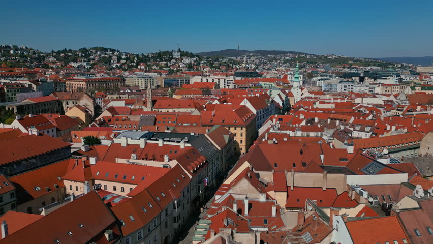 An aerial view shows Bratislava old town with a density of red-tiled roofs, leading to distant green hills under a clear blue sky in Slovakia
