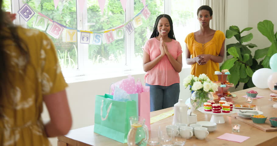 Diverse female friends clapping at baby shower table, welcoming expectant mother, draping sash. Celebration, friendship, joy, hospitality, decor, floral, merriment