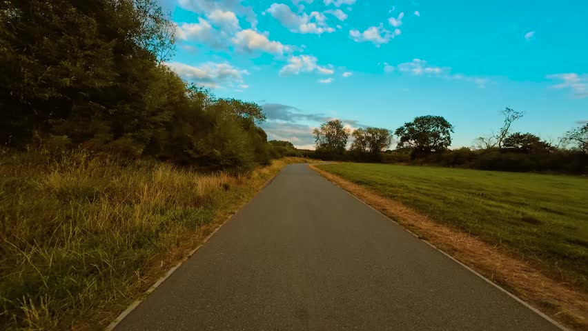 Biker riding bicycle on curving asphalt path through green fields and trees under blue sky POV. Cyclist cycling along countryside road surrounded by grass and bushes with scattered clouds first person