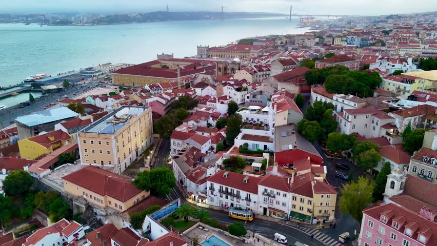 A drone footage of the historic yellow tram 28 passing through the narrow streets of Alfama district near the Tagus River in Lisbon, Portugal.