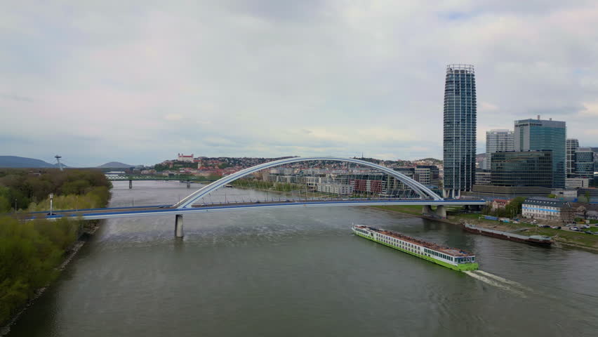 A drone view of a river cruise ship passes in the Danube River under the Apollo Bridge between urban and wooded banks in Bratislava, Slovakia