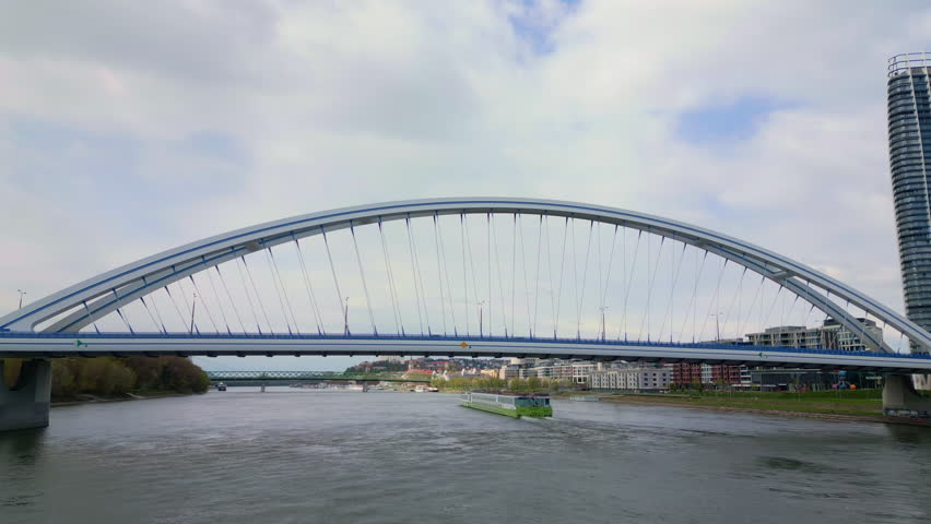 A drone view of a river cruise ship passes in the Danube River behind the Apollo Bridge between urban and wooded banks in Bratislava, Slovakia