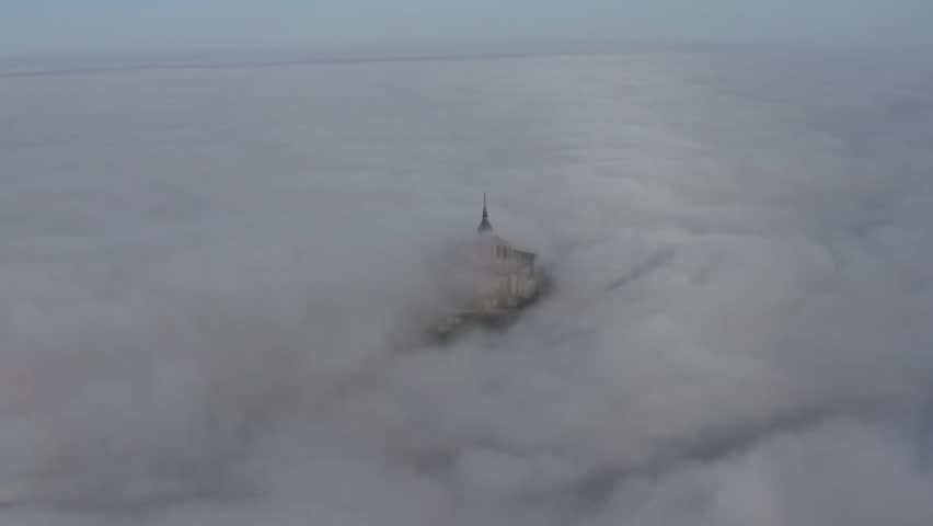 Aerial view of Mont Saint-Michel piercing through a sea of dense fog, its silhouette a stark contrast against the soft, ethereal clouds, Mont Saint-Michel, Normandy, France.