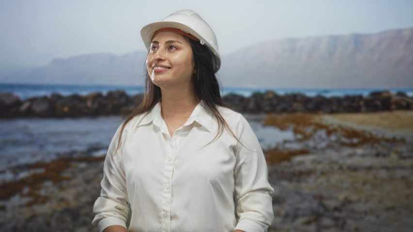 Woman wearing hard hat smiles and looks out across rocky shoreline on beach; coastal professional optimism.