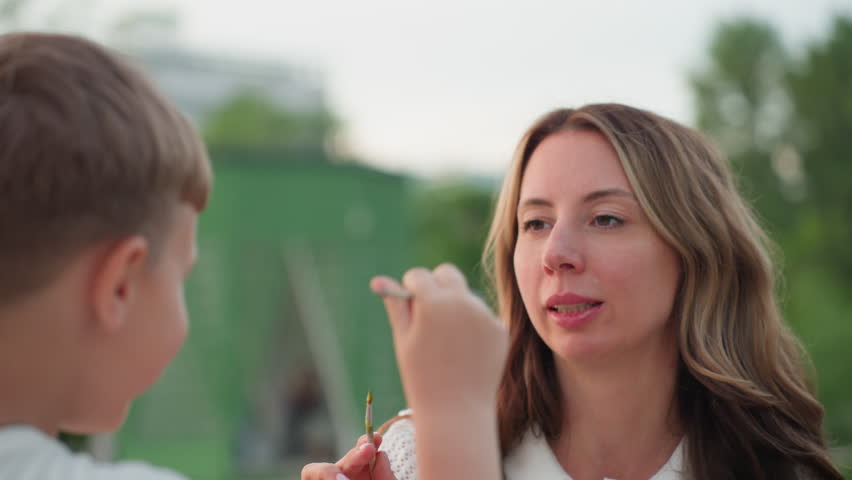 close up of child hand waving paint brush toward mom while seated on wooden dock by pool under sunset glow, capturing gentle playful shared art moment with water reflection and warm evening light