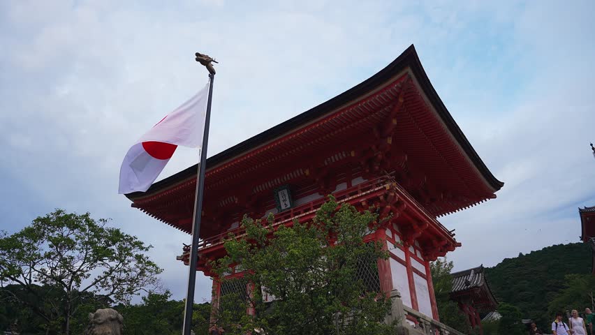A vermilion two story shrine gate in Kyoto, Japan, stands with a flag as visitors walk and birds move. Trees and hills frame the scene under pale cloudy light.