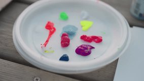 hand holds brush mixing pastel blue acrylic with neon red green yellow pink purple paints on white plastic palette plate on wooden bench by river at sunset outdoor painting session - Powered by Shutterstock - Get 15% off with code: PIKWIZARD15