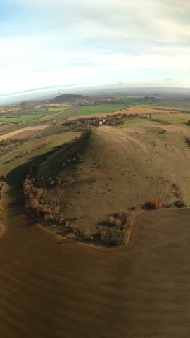 A vertical view of the Cicov Peak in the Czech Republic, showing its rounded volcanic hill rising above autumn fields under daylight and open skies