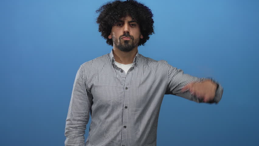 Man with curly hair saluting with hand to forehead in studio against solid blue background wearing checkered shirt; confidence determination focus.