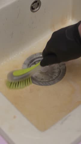 Person scrubs stained kitchen sink with brush and glove as grime begins to lift, revealing cleaner surface beneath heavy buildup.