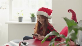 A cheerful woman in a festive Santa hat happily and enthusiastically uses her smartphone in a cozy, beautifully decorated living room, creating a delightful and joyful Christmas atmosphere all around - Powered by Shutterstock - Get 15% off with code: PIKWIZARD15