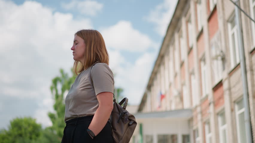Sad young woman with backpack standing outdoors near building under bright sky, bringing out napkin from pocket to wipe tears, emotional expression showing sorrow