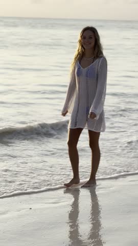 A young woman in a white dress walks along a pristine beach at sunrise with waves gently touching the shore. Perfect for relaxation, travel, and mindfulness themes.