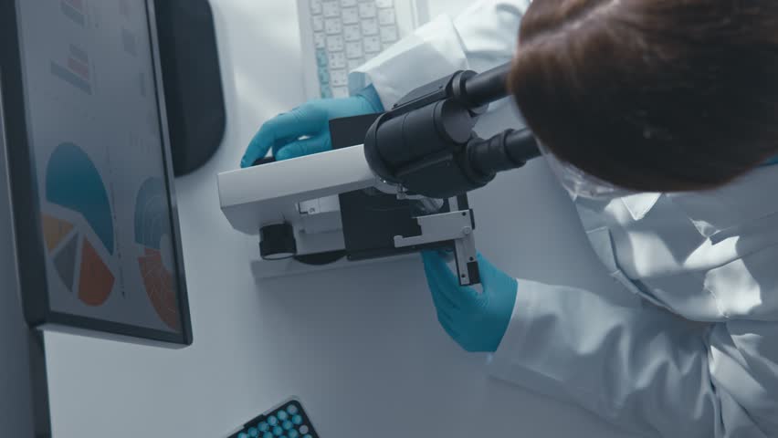 A scientist wearing gloves executes detailed microscopic analysis, documenting findings on a computer while utilizing advanced lab equipment in a clean, modern research environment.