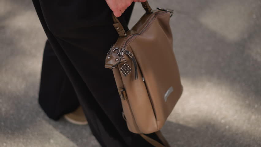 Top down view of woman walking under bright sunlight holding brown leather bag, moving toward building, soft blurred background with shadows on pavement