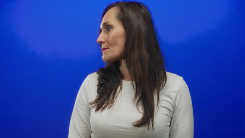 Woman with long hair wearing white shirt standing against vibrant blue background wall, gazing sideways, in a calm and contemplative pose.