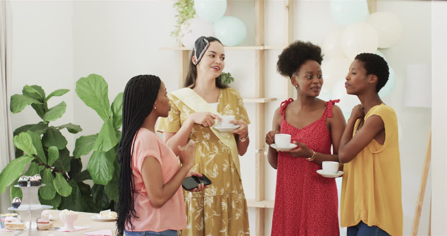 Diverse female friends at baby shower celebrating holding teacup touching bump taking phone selfie. Celebration, friendship, female, elegance, joy, leisure, decor