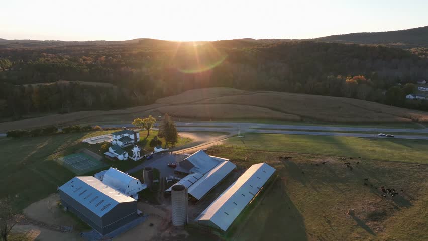 Aerial rising shot of american farmstead with barn and buildings in USA. Sunset light behind green mountains of Lynchburg, Virginia. Rural area of american town with road.