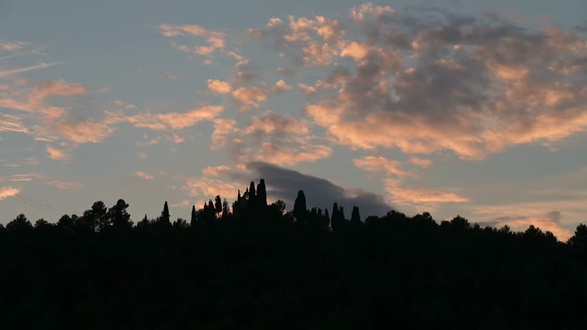 Pink and grey clouds glide over the sky above a shadowy forest outline as the sun sets. Gradually, the sky transitions from evening light to complete darkness