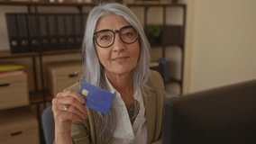 Woman smiling in an office holding a blue credit card, showcasing senior professional lifestyle with a confident expression and grey hair in work environment - Powered by Shutterstock - Get 15% off with code: PIKWIZARD15